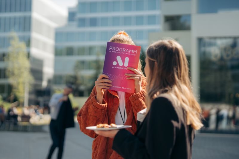 Zwei Besucherinnen im Gespräch vor dem Programmheft der AI in Marketing Konferenz